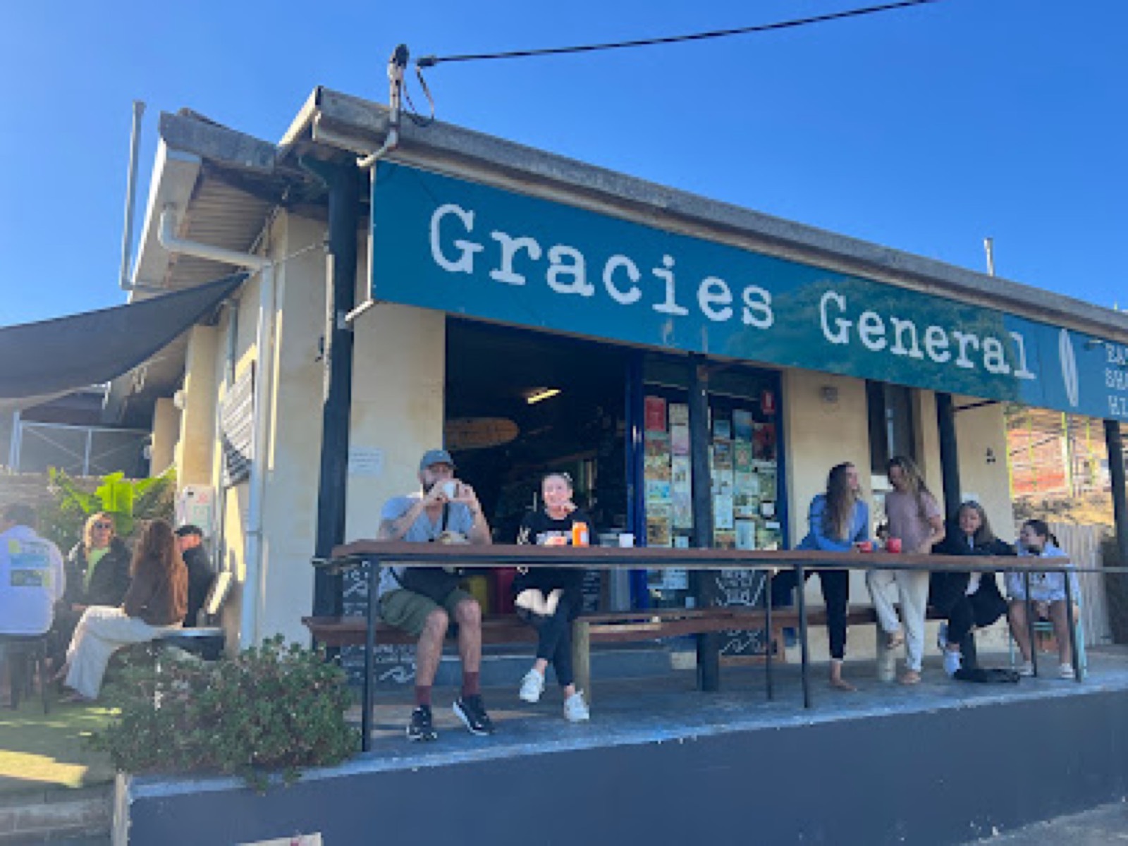The blue Gracies General storefront with people relaxing at picnic benches out front and a surfboard sign, on a sunny day in Gracetown